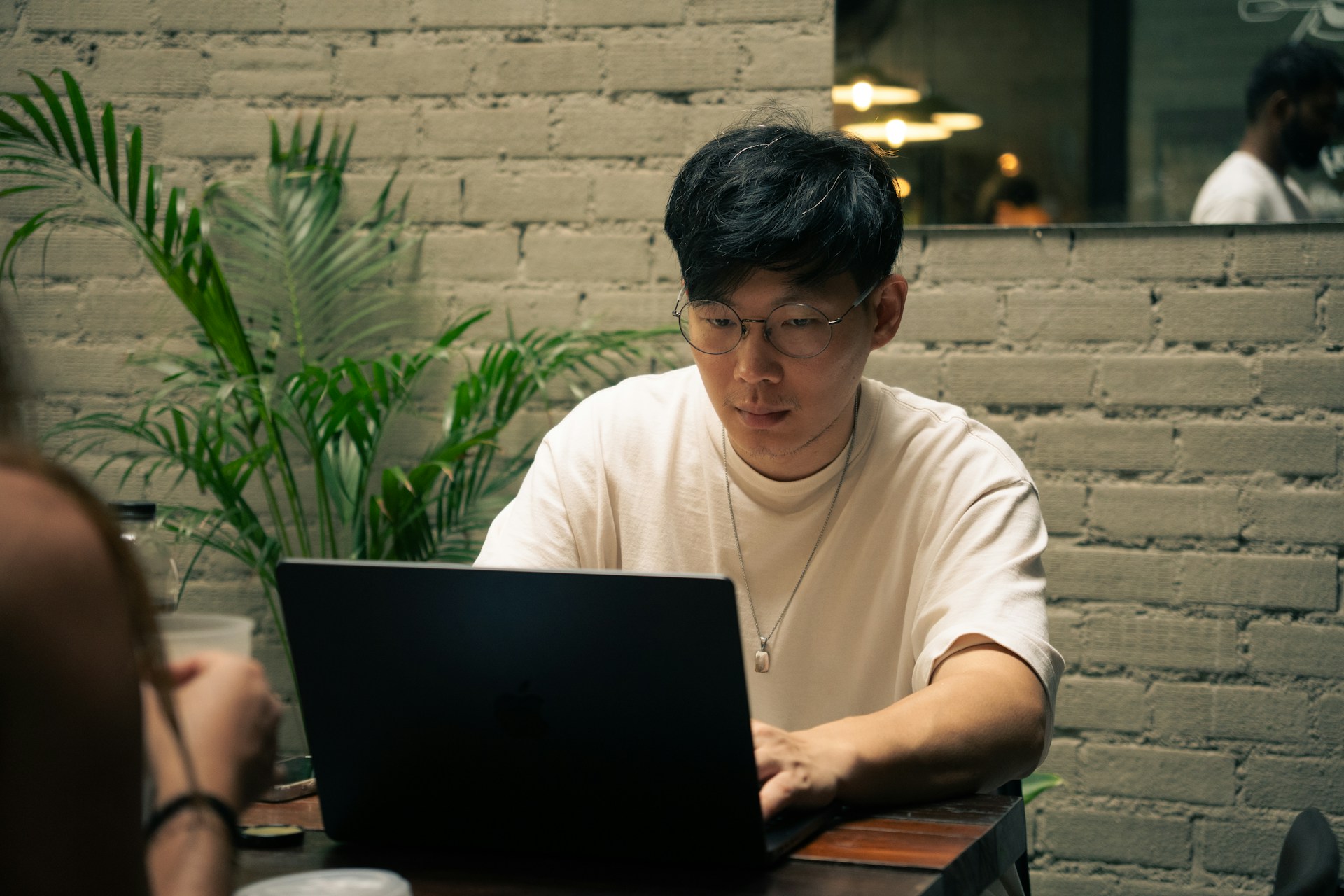 レンガの壁とグリーンを背景に、カフェでノートパソコンに向かって集中して作業する男性。